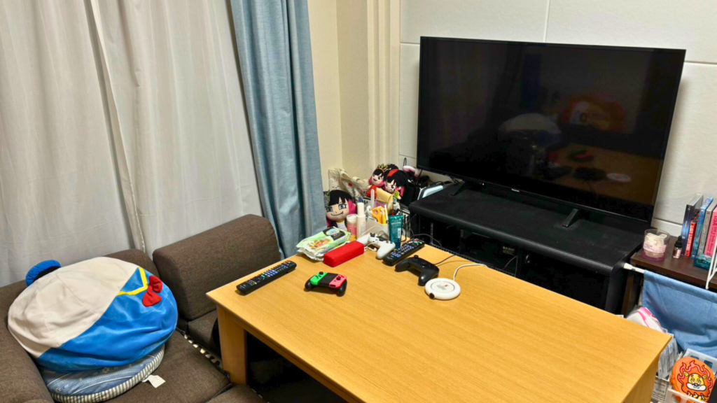 女性の部屋で、テレビの前にローテーブル、その上にはリモコンやゲーム機、ぬいぐるみなど置かれていて、さらに手前にソファとドナルドダックのクッションが配置されている