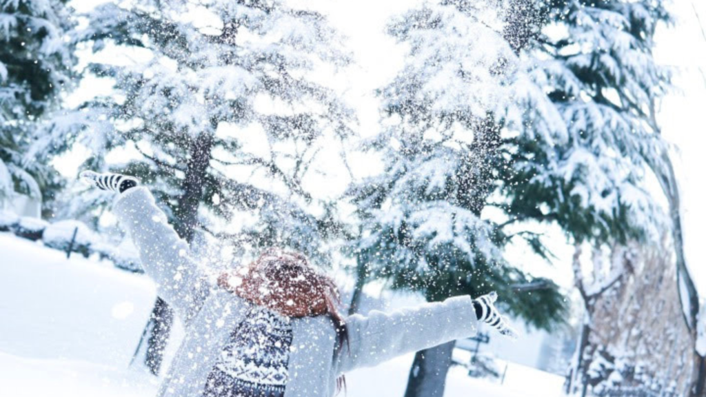 針葉樹の広がる雪景色の中、赤いマフラーにグレーのコート、ボーダー柄の手袋にセーターの女性が雪を空に向かって両手で投げ広げている