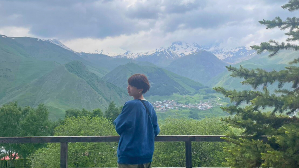 海外の壮大な雪山が奥に広がる中、手前は青々とした山に囲まれた小さな街があり、それを展望台から見つめながら振り返る青いシャツにショートヘアの女性