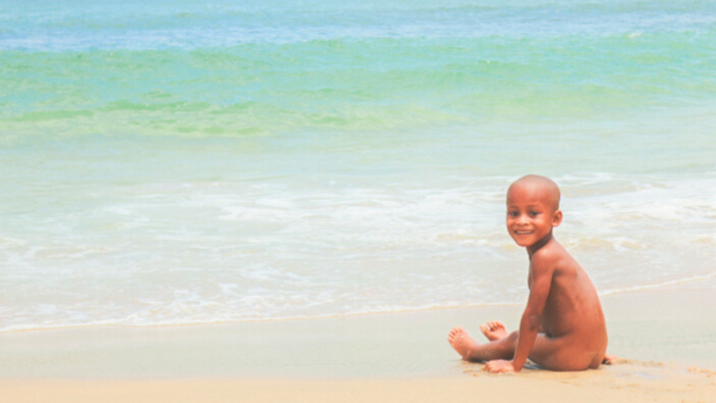 透き通る青い海の砂浜で、裸で座りながらこちらを振り返るアフリカ系の男の子。