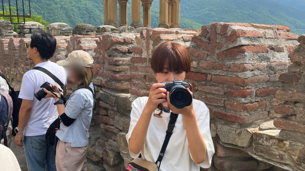 大自然の中のレンガ調の建造物の中で白いTシャツにショートヘアの女性が一眼レフをこちらに向けてレンズを覗いている
